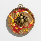 A round glass pendant featuring a metallic gold bee motif over a marbled background of brown, red, and yellow, with a gold hanging loop at the top.