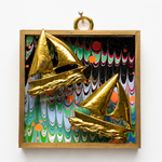 A framed wall art piece featuring two metallic gold 3D sailboats set against a colorful marbled background with green, blue, pink, and black patterns, enclosed in a light wood frame.